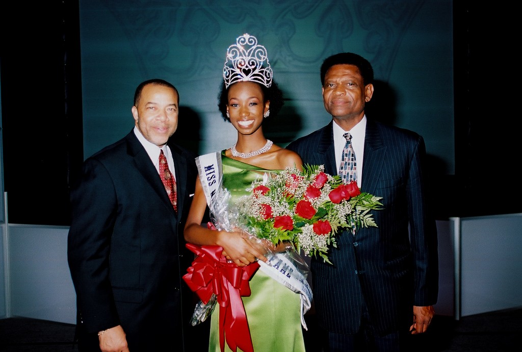 A crowned queen holding roses on stage with two men in formal attire